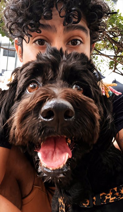 A close-up selfie of a young person with curly hair posing behind a fluffy black dog.