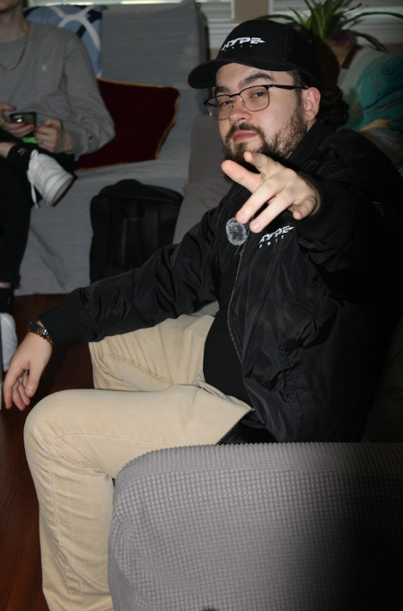 A bearded man wearing glasses, a black cap with "FTP" logo, and a black jacket sits on a gray couch, gesturing toward the camera. Another person is partially visible on the left holding a camera.
