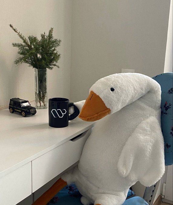 The image features a large white stuffed animal duck sitting on top of a desk next to a black coffee mug with a white "W" logo, as well as a toy car. The scene appears to be set up for a photo session or a social media post related to DeFi and the concept of real utility in the crypto world.