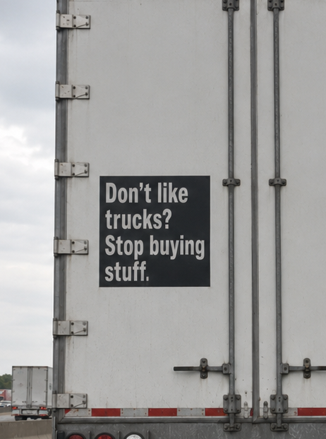 The back of a white semi-truck trailer displays a black sign with white text reading 'Don't like trucks? Stop buying stuff.'