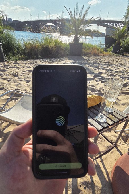 A hand holds an iPhone displaying a fingerprint icon and "Unlock" prompt from the Jupiter Mobile app on a sunny beach with palm trees and a bridge in the background. The image showcases the app's biometric login feature in a scenic riverside setting.