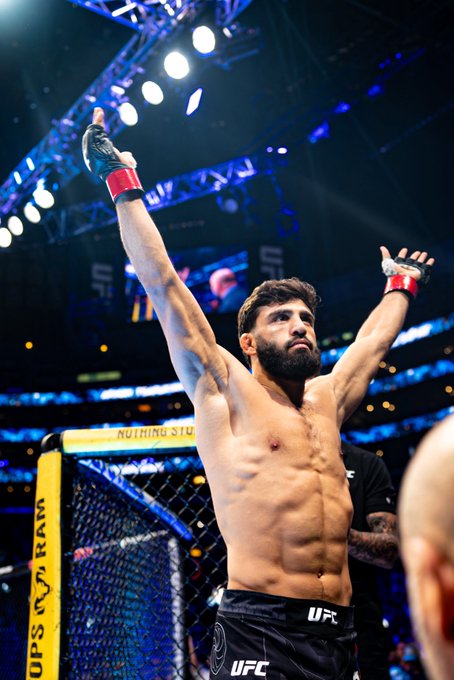 A bearded, shirtless male UFC fighter wearing black "UFC" branded shorts and red-taped MMA gloves raises both arms triumphantly inside the octagon at the T-Mobile Arena during UFC 331, with the "Crypto.com" sponsorship banner clearly visible on the cage padding behind him and arena spotlights glowing above, perfectly complementing the post's promotion of Crypto.com's partnership with the UFC event now streaming live on Paramount+.