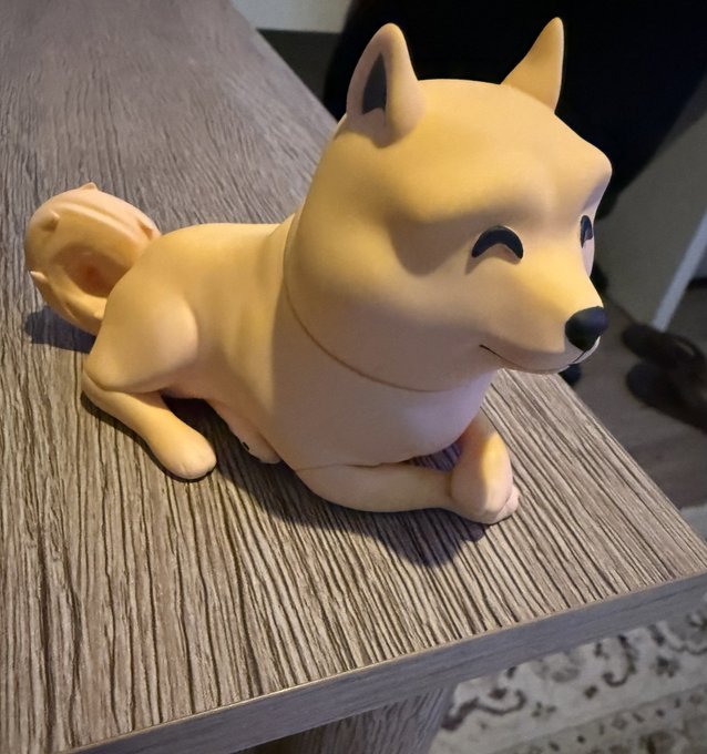 The image features a small, light brown dog figurine sitting on top of a wooden table. The figurine appears to be made out of plastic or ceramic material, giving it a cute and playful appearance. In the background, there are two people present - one person is standing near the left side of the frame, while another person can be seen in the right corner of the image.

The post that accompanies this image reads: "My Son knows me well #shibainu #Bullrun incoming". This message seems to be a reference to the dog figurine sitting on the table and the two people present in the background, suggesting that the person who posted the image might have a close relationship with their son or someone else named Bullrun.
