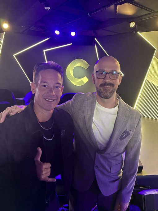 Two men smiling and posing together at an event with purple lighting; the man on the left wears a black jacket, while the man on the right wears glasses and a grey plaid blazer, with a large 'C' logo visible on the wall behind them.