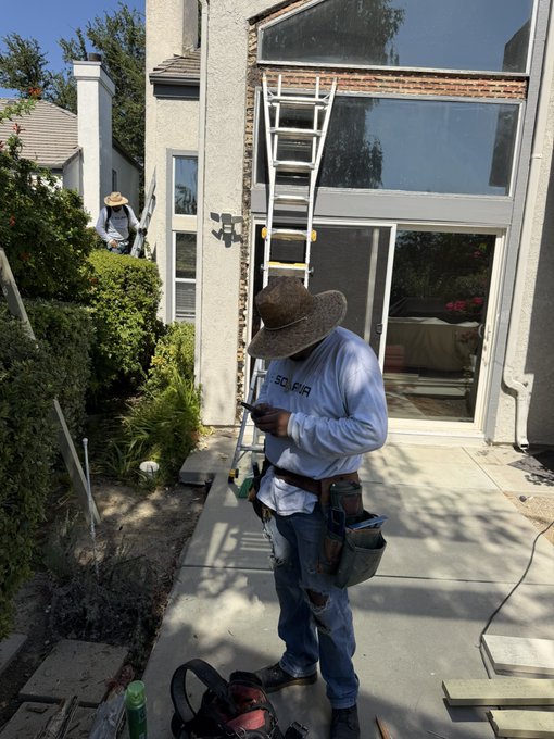 A construction worker wearing a wide-brimmed hat and a tool belt stands in a backyard looking at his phone, with a tall aluminum extension ladder propped against the second story of the house.