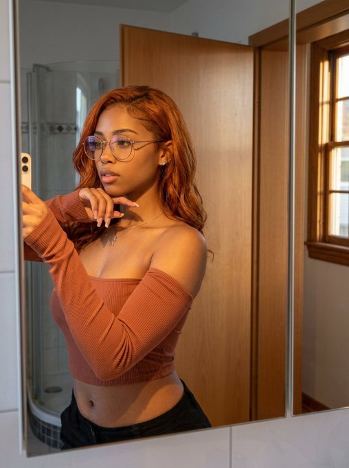 A mirror selfie of a young woman with long reddish-brown hair and glasses, wearing a brown off-the-shoulder top, posing in what appears to be a bathroom.