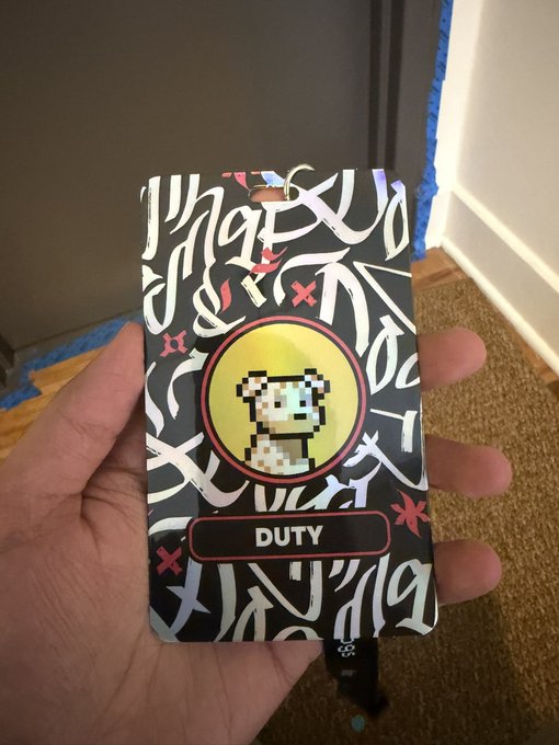 A hand holds up a holographic trading card featuring a pixelated bear character labeled "DUTY" on a black background with graffiti-style lettering.