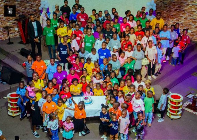 The image shows a large group of children gathered together for a photo, with many of them wearing colorful shirts. The background features a stage or podium set up, suggesting that this gathering might be part of an event or celebration. There are also speakers present in the scene, indicating that there could be announcements or speeches taking place during the event.
