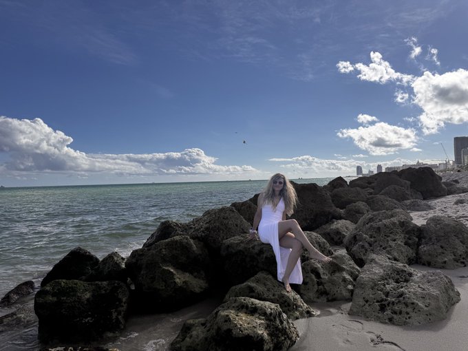A woman with long blonde hair in a white dress sits on large rocks along a shoreline, with a city skyline visible in the distance under a partly cloudy sky.