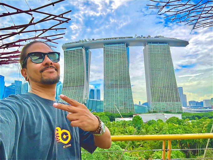 A man in sunglasses and a gray "JC CANE" t-shirt poses with a peace sign in front of the iconic Marina Bay Sands hotel in Singapore, matching the cheerful "GM" (Good Morning) vibe with a sunny, optimistic travel shot.