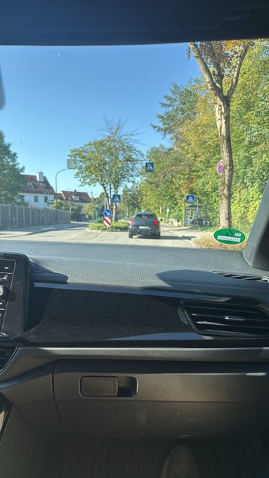 View from inside a car looking through the windshield at a sunny street scene with trees, road signs, and a parked car ahead.