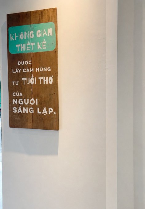 A wooden sign with white text on a teal accent reads "KHÔNG GIAN THIẾT KẾ ĐƯỢC LẤY CẢM HỨNG TỪ TUỔI THƠ CỦA NGƯỜI SÁNG LẬP" (Translation: "The design space is inspired by the childhood of the founder"), hanging on a white wall.

The Vietnamese signage about childhood inspiration reflects the post's message of steady, personal growth—reminiscent of drawing from one's own roots and simple dreams (like receiving a $1xxx mail) rather than competing with others.