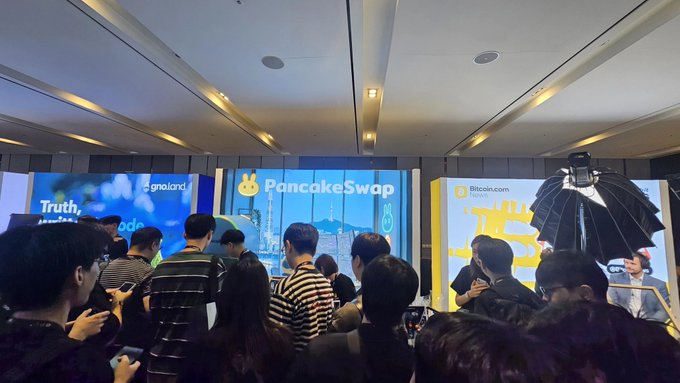 A crowd of people attends a conference with screens displaying logos including 'PancakeSwap' and 'Bitcoin.com Token', suggesting a crypto industry event.