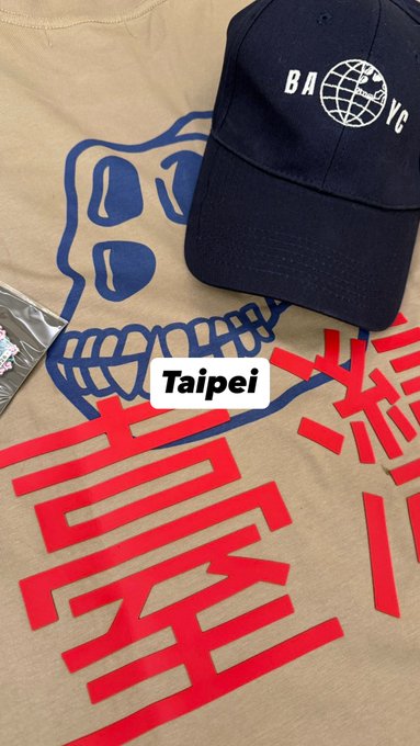 A navy BAYC (Bored Ape Yacht Club) baseball cap rests atop a tan t-shirt featuring a blue skull graphic and red Chinese characters, with a "Taipei" label indicating the location. The image showcases Taipei's tour merchandise as part of the BAYC x Asia tour merch comparison.