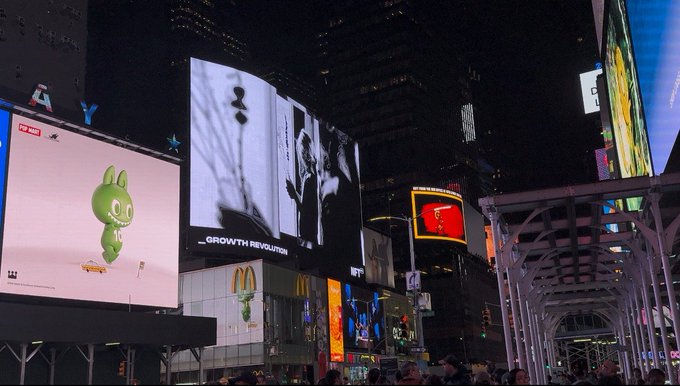 A nighttime view of Times Square in New York City, featuring illuminated billboards including an 'iQIYI' ad with a green cartoon character, and a McDonald's sign.