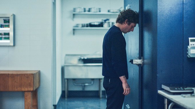 The image shows a man standing in front of an open blue door, leaning against it with his head down. The room appears to be a kitchen or dining area, as there are several bowls placed around the space. A wooden table can also be seen nearby. The scene seems to depict a moment of contemplation or sadness for the man, possibly due to a recent transaction involving cryptocurrency.
