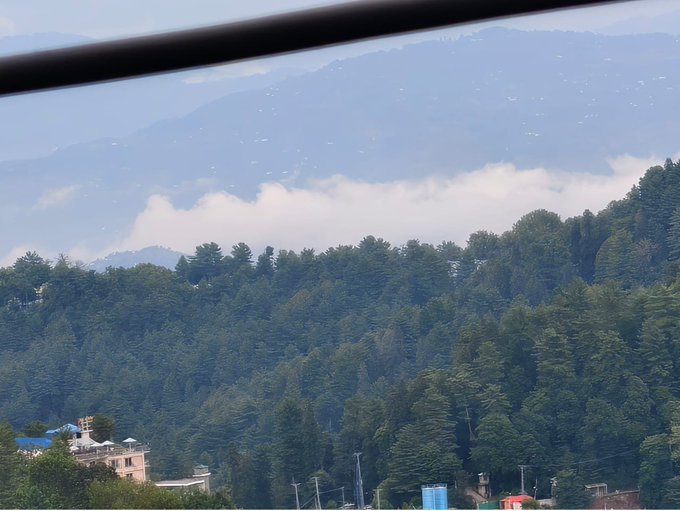 A scenic mountain landscape view through a window, showing lush green pine-covered hills with misty clouds and distant mountain peaks under a blue sky.