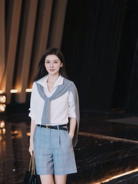 A young woman poses in a white blouse with a blue sweater draped over her shoulders and light blue shorts, holding a handbag in a softly lit indoor setting.