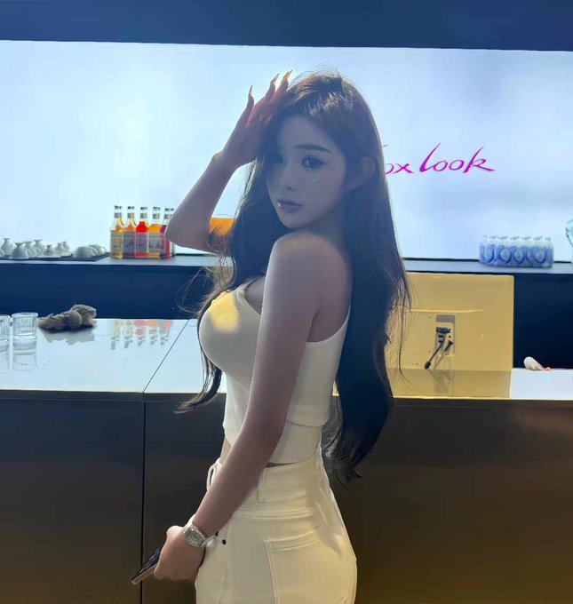 The image features a woman with long hair wearing white clothing, standing next to a bar counter filled with various bottles of alcohol. She has her hand up to her head, possibly posing for a photo or engaging in conversation at the bar. The background includes a TV screen and other people present but not directly involved with the main subject.