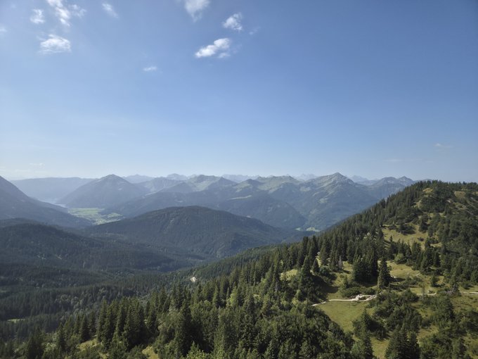 A scenic mountain landscape view with rolling green hills, dense forests, and a clear blue sky with scattered clouds.