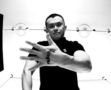 A black-and-white photo of a young man in a polo shirt extending his hand toward the camera, with badminton rackets visible on a rack behind him.