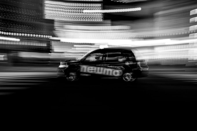 A black-and-white motion-blurred photograph of a small hatchback car with 'newmo' branding on the side, speeding through a city at night.