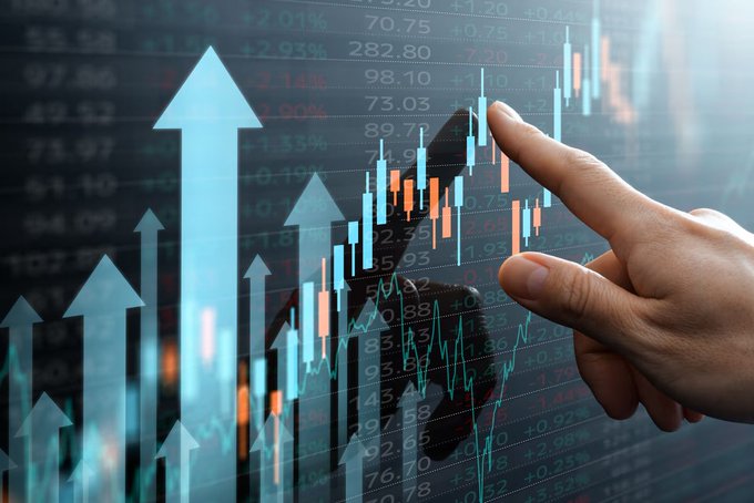 A hand pointing at a financial data display showing stock market candlestick charts, upward arrows, and various numerical percentages indicating gains and losses.