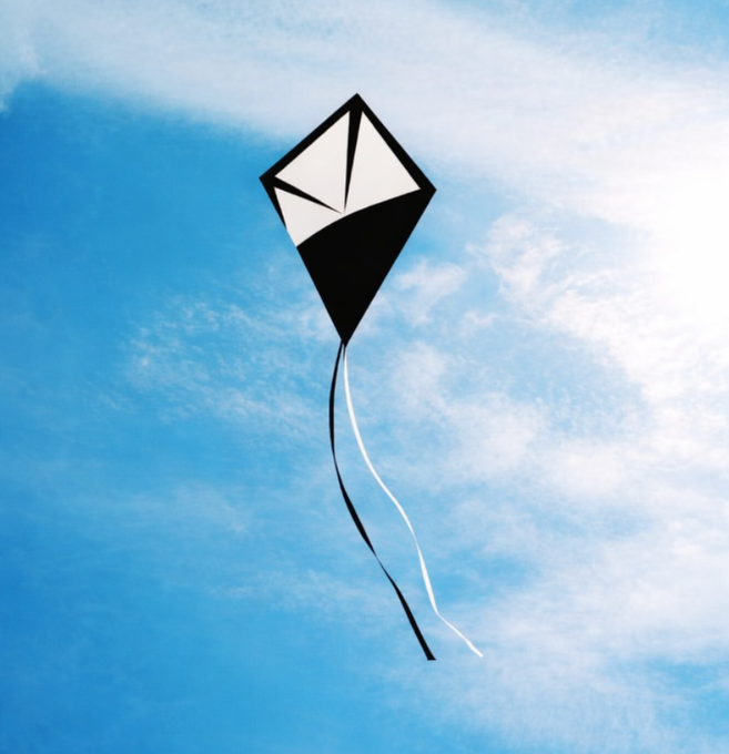 A black and white diamond-shaped kite flies against a bright blue sky with wispy clouds, featuring two long ribbon tails trailing below it.