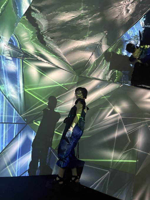 A person wearing dark sunglasses, a dark vest, and blue pants stands against a wall projected with green geometric lines and light patterns, casting a distinct shadow.