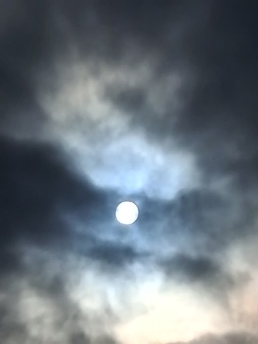 A photograph of a bright full moon glowing through dark, wispy clouds in a night sky.