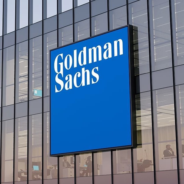 A large illuminated "Goldman Sachs" sign is mounted on the glass facade of an office building, with workers visible through the windows below. The image visually represents the investment bank mentioned in the news post warning of potential $185 billion in forced global market selling this month.
