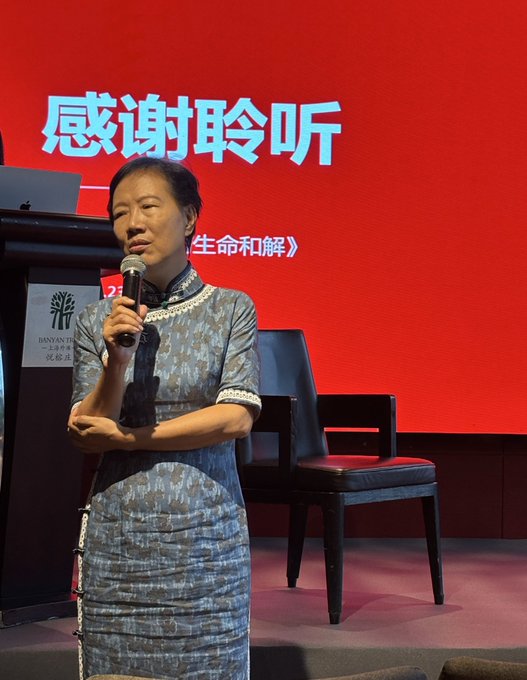A woman in a grey traditional Chinese dress (qipao) speaking into a microphone on stage with a red screen behind her displaying the Chinese text '感谢聆听' (Thank you for listening).