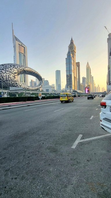 A street view of Dubai featuring the Museum of the Future with its distinctive Arabic calligraphy-covered torus shape, alongside the city's skyline including tall skyscrapers at sunset.