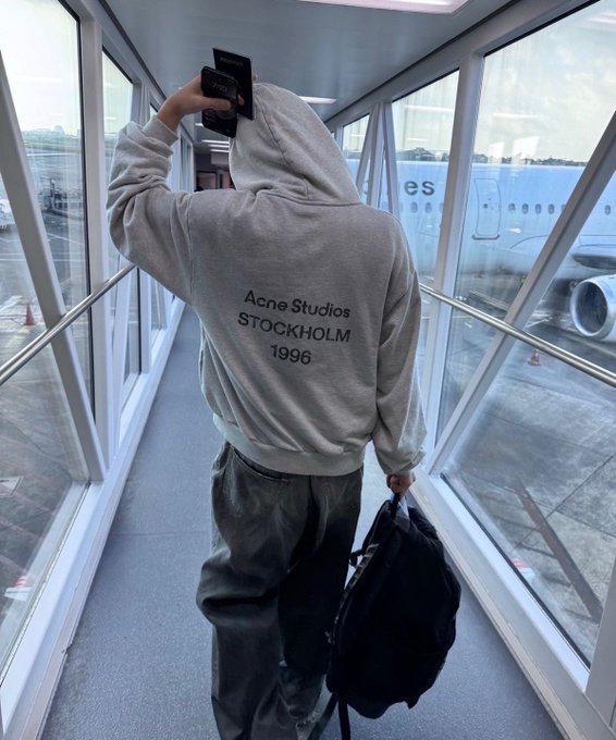 A person wearing a beige Acne Studios Stockholm 1996 hoodie stands in an airport jet bridge holding up a passport and a black duffel bag, with an airplane visible outside the window.