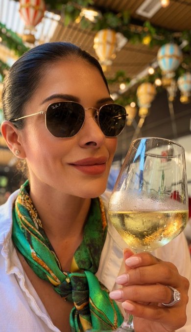 A stylish woman in sunglasses and a vibrant green patterned scarf smiles while holding up a glass of white wine at an outdoor venue decorated with festive hanging lanterns, perfectly capturing the cheerful "drinking my feelings" sentiment with her elegant toast.