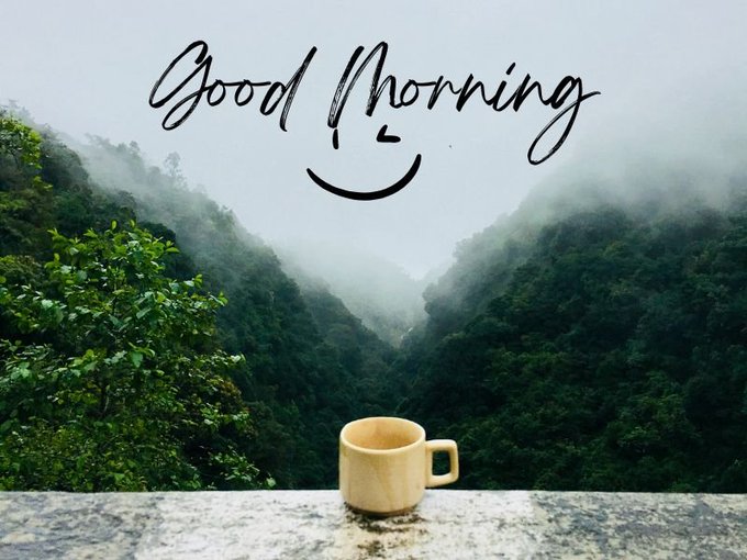 A beige mug sits on a ledge overlooking a misty, green mountain valley, with 'Good Morning' written in cursive above a hand-drawn smiley face.