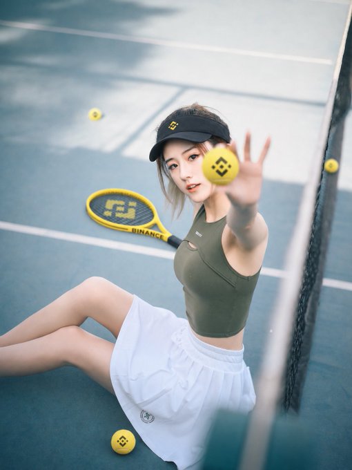 The image features a woman sitting on the ground with her legs crossed, holding a tennis racket and several tennis balls around her. The background of the photo shows a tennis court with a net dividing it into two halves. There are also some yellow tennis balls scattered in the scene. The caption to the image reads "#币安周边大使". This suggests that there might be an event or gathering related to tennis, possibly involving a competition or exhibition match.