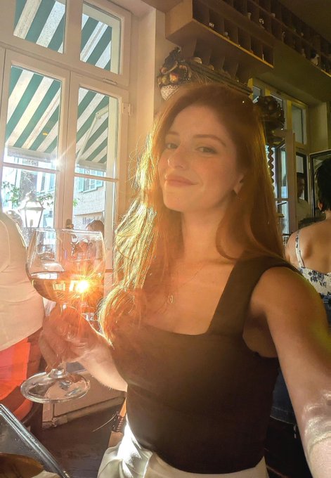 A woman with long reddish-brown hair smiles softly while holding a glass of rosé wine in a sunlit restaurant or wine bar. The image relates to the post by evoking a relaxed, celebratory atmosphere — fitting for the question of whether the crypto bull market has officially started, suggesting a moment of optimism and toasting to potential gains.