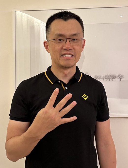 A man wearing glasses and a black polo shirt with a yellow logo smiles while holding up four fingers, with a framed black-and-white landscape photo visible behind him.
