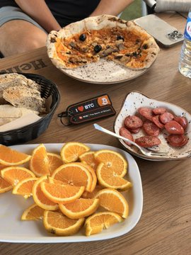 A meal spread on a wooden table featuring a slice of pizza, a basket of bread, a bowl of sliced sausages, and a plate of orange slices.