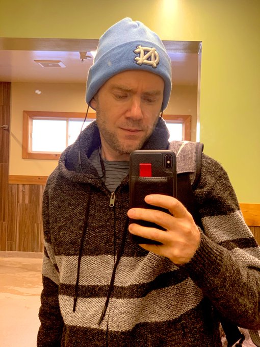 A man wearing a blue North Dakota beanie and a striped hoodie is taking a mirror selfie with his smartphone in a wood-paneled room, likely sharing the news about the upcoming XNT airdrop to the X1 community.