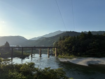 A scenic photograph of a bridge spanning a river with forested mountains in the background under a clear blue sky, taken from a slightly elevated vantage point.