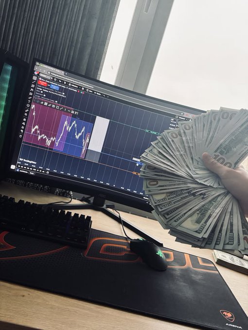 A photograph of a computer monitor displaying financial charts and trading software, next to a keyboard, gaming mouse on an 'Arena' mousepad, and a hand holding a fan of US dollar bills.