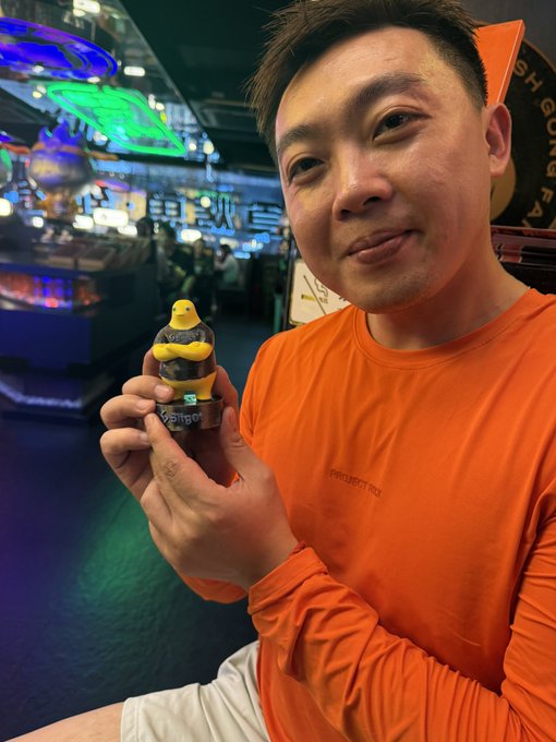 A man in an orange shirt smiling while holding a chocolate lava cake dessert topped with a yellow character figure in a brightly lit arcade or gaming venue.