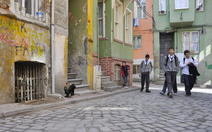 Four young men wearing school uniforms and backpacks walk down a cobblestone street in an urban neighborhood with colorful, weathered buildings, while a black cat sits on the sidewalk.