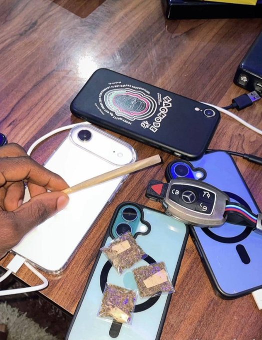 A hand holds a pre-roll joint over a table covered with several smartphones, a phone case with a brain graphic, a car key fob, and small plastic bags containing what appears to be cannabis.