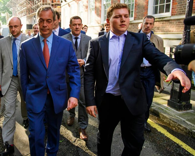 Nigel Farage, dressed in a blue suit with a red tie, walks alongside a younger man in a dark suit and lavender shirt, surrounded by a group of associates and security personnel on a city street. The image illustrates the post's focus on Farage, who is under scrutiny for reportedly receiving support from George Cottrell—a convicted fraudster now linked to a crypto casino—before entering Parliament, raising concerns about his ties to the crypto industry.