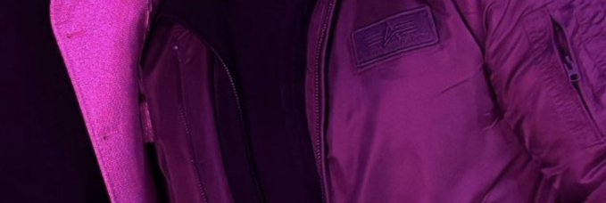 The image shows a purple jacket with a black zipper, partially open revealing its interior lining. The jacket has an "Alpha Alert." label attached to the back pocket. The background of the image is a light gray color that contrasts with the vibrant purple of the jacket and the pinkish hue of the zipper.