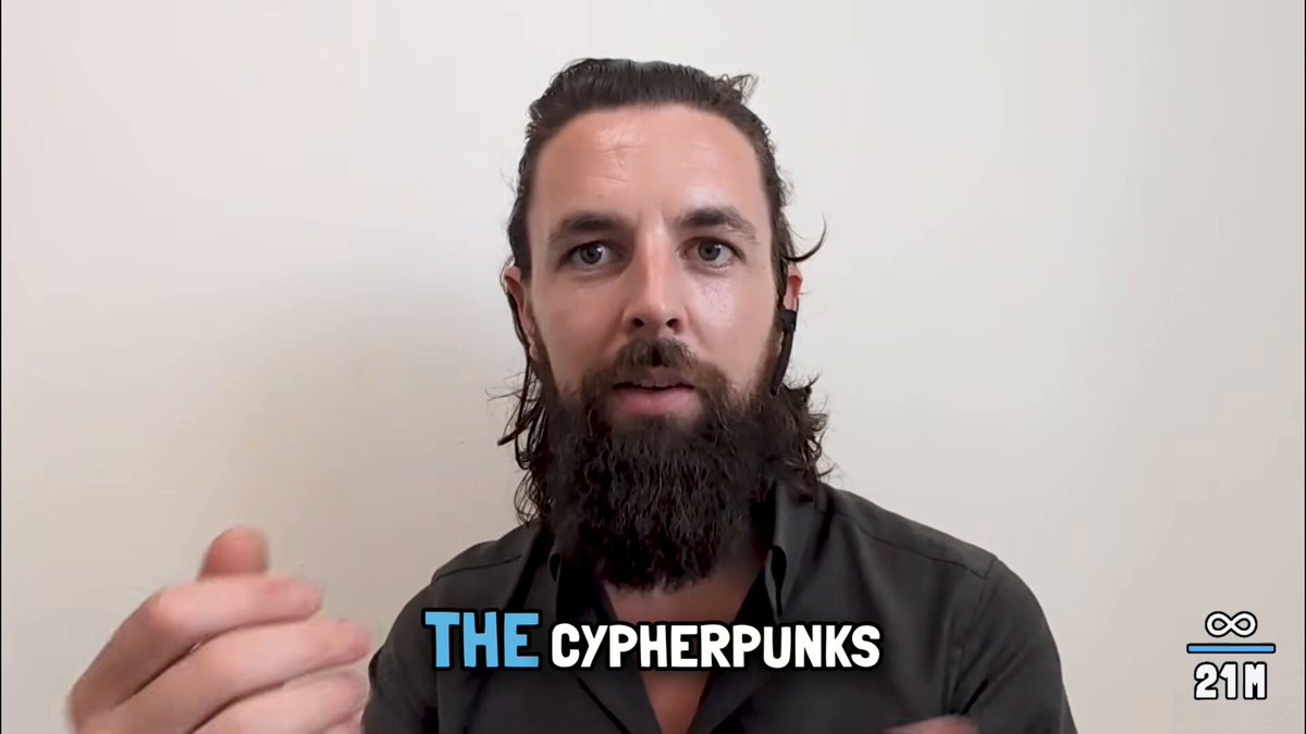 The image features a man wearing a black shirt with white text that reads "The Cyperpunks". The man has a beard and mustache, giving him a hipster appearance. He appears to be talking or posing for a photo, possibly at an event related to the "Cyberpunk" subculture.