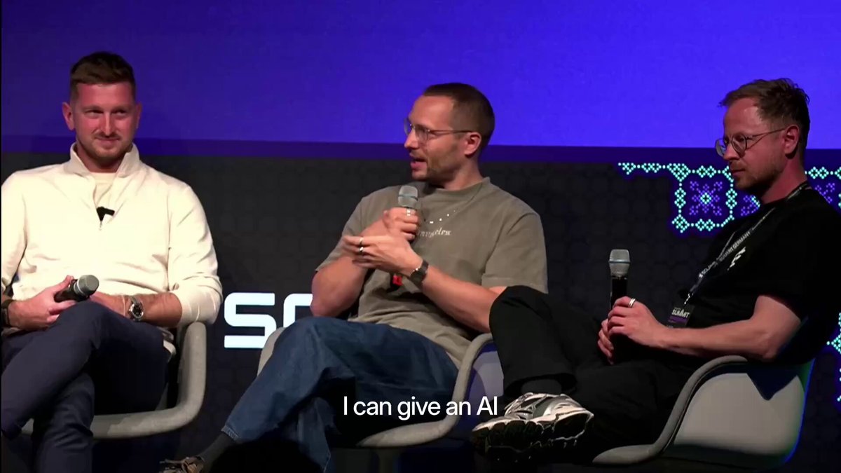 The image shows three men sitting down, with one man holding a microphone while another two are seated behind him. They appear to be engaged in conversation or discussion. In the background, there is a large screen displaying text that reads "I can give an AI". The focus of this image seems to be on the interaction between these individuals and their surroundings.